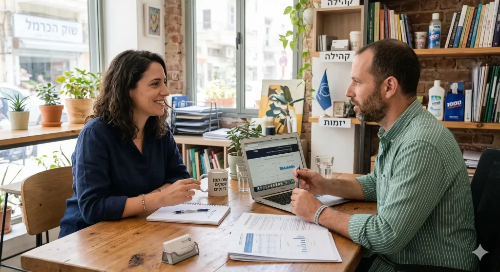 שני אנשים נפגשים במשרד לשיחה עסקית, עם מחשב נייד ומסמכים על השולחן, בסביבה מוארת ונעימה.
